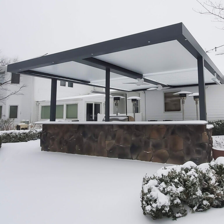 Large dark-framed aluminum louvered pergola with closed louvers, ceiling fan, recessed lighting, and freestanding heaters over a stone bar counter in a fully snow-covered backyard