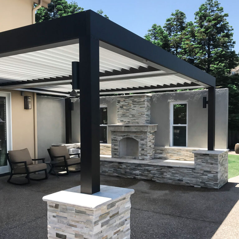 Dark-framed freestanding aluminum louvered pergola with open white louvers and stone pier bases beside a large stacked stone outdoor fireplace, with ceiling fan and lounge chairs on a concrete patio