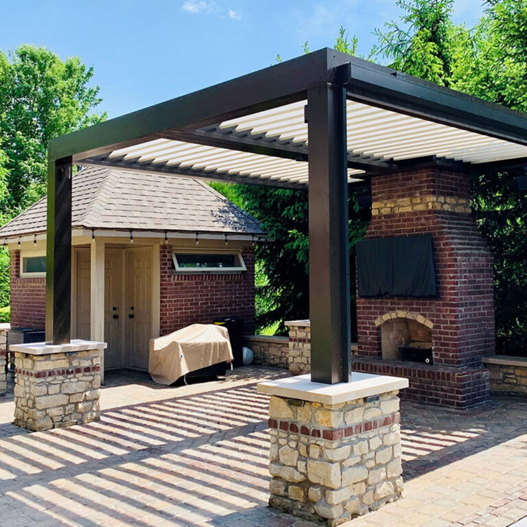 Dark-framed freestanding aluminum louvered pergola with open cream louvers on stone pier bases on a brick paver patio, beside a brick arched fireplace with outdoor TV and a pool house in the background