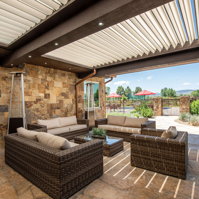 Under-canopy view of a dark-framed aluminum louvered pergola with open louvers, recessed downlights, and two freestanding tower heaters over a wicker lounge on a flagstone patio beside a natural stone wall, with a mountain view beyond