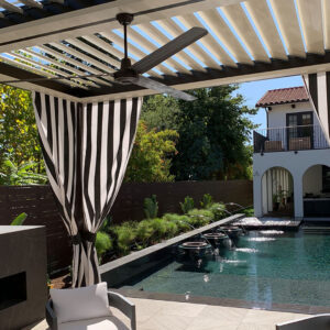 Under-canopy view of a dark aluminum louvered pergola with open cream louvers, wood-blade ceiling fan, and black-and-white striped curtains beside a dark-tiled pool with fountain jets and a Spanish Colonial home beyond