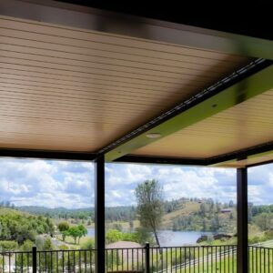 Underside of a closed dark-framed aluminum louvered pergola with recessed speakers, looking out over a lake, rolling hills, and open sky from an elevated deck