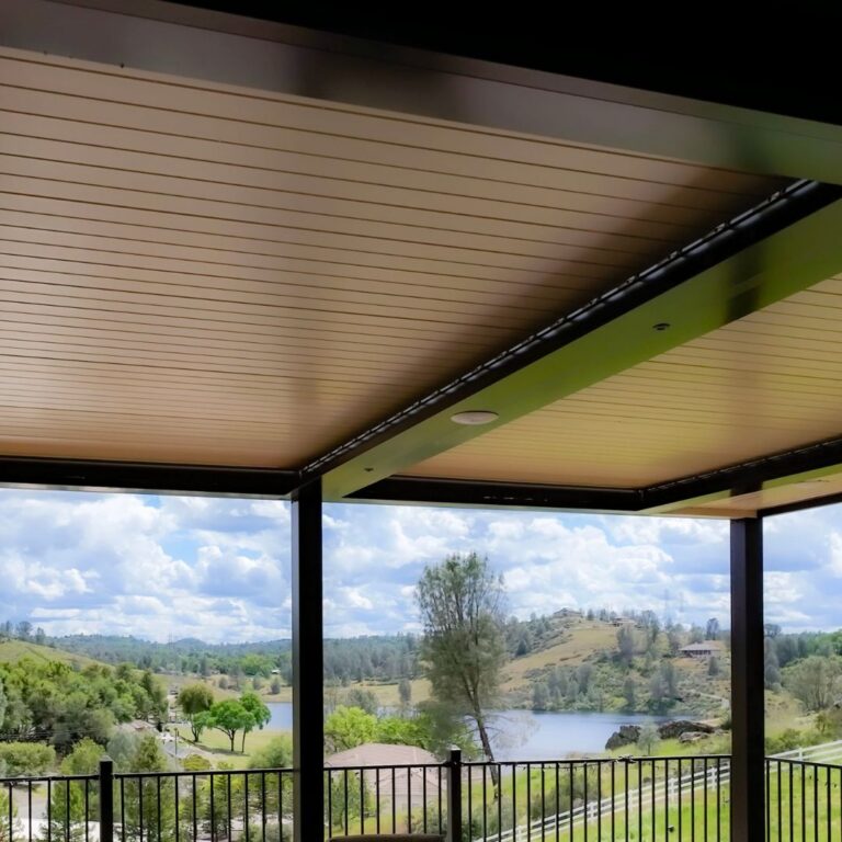 Underside of a closed dark-framed aluminum louvered pergola with recessed speakers, looking out over a lake, rolling hills, and open sky from an elevated deck