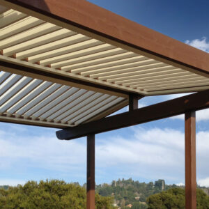 Low-angle view of an aluminum louvered pergola with a wood-grain finish frame and open cream louvers against a blue sky with a treetop and rolling hillside below