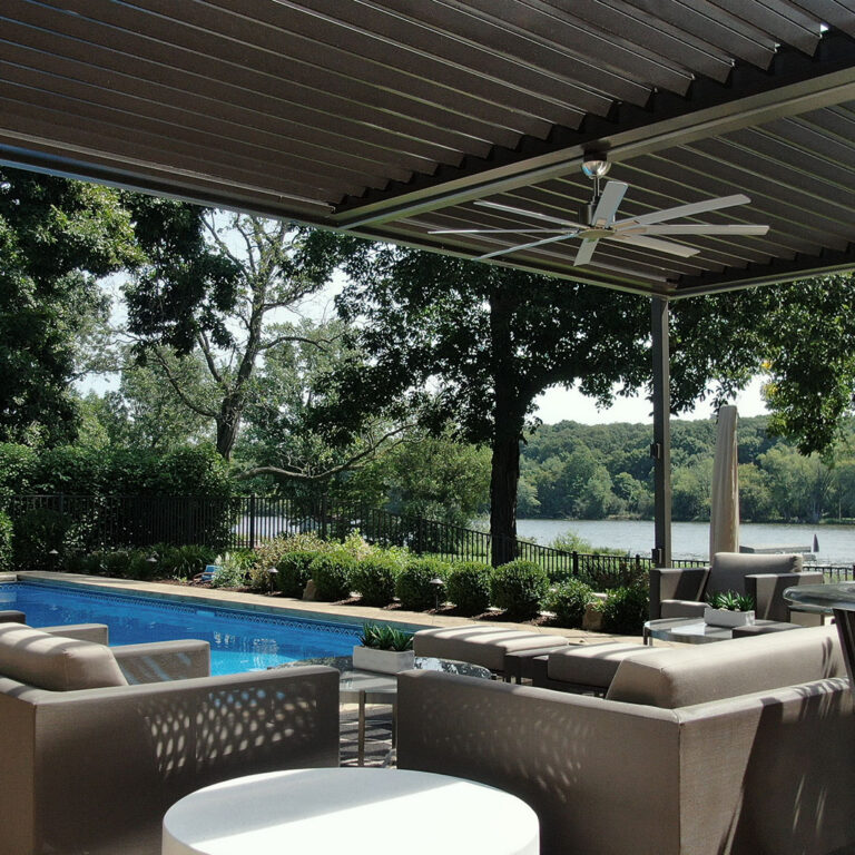 Under-view of a dark aluminum louvered roof with a ceiling fan mounted above poolside seating, overlooking a lake through mature trees