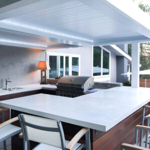 White aluminum louvered roof pergola with closed louvers and integrated LED strip lighting illuminating a modern outdoor kitchen with built-in grill and bar seating at dusk