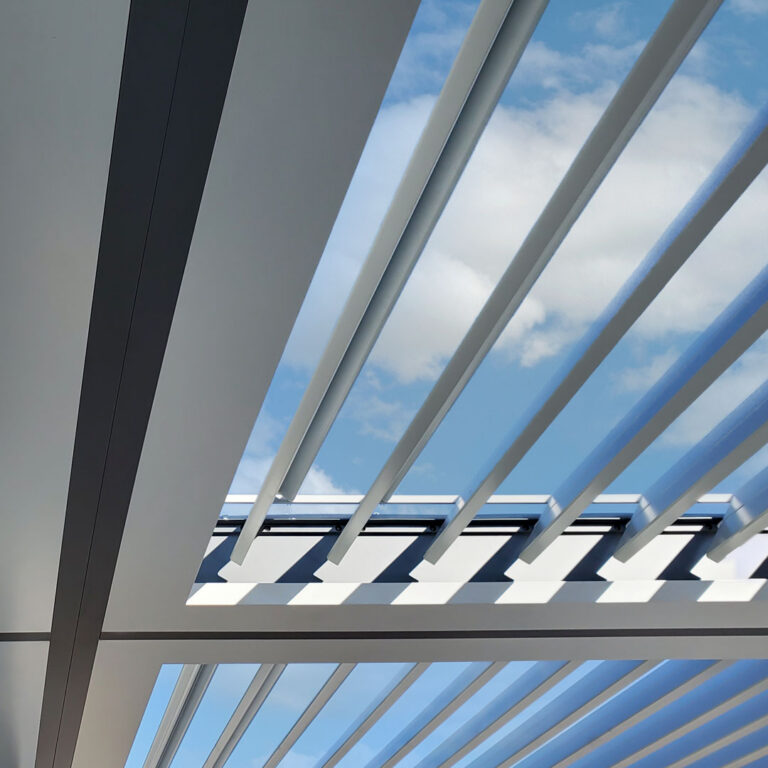 Close-up upward view of open white aluminum louvers on a pergola showing the louver pivot and drive mechanism hardware against a blue sky with clouds