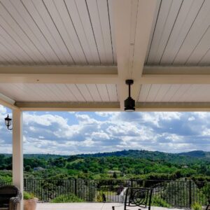 View from beneath a closed white aluminum louvered roof with a compact ceiling fan mounted at center, overlooking a wide valley and rolling hills beyond an iron-railed deck