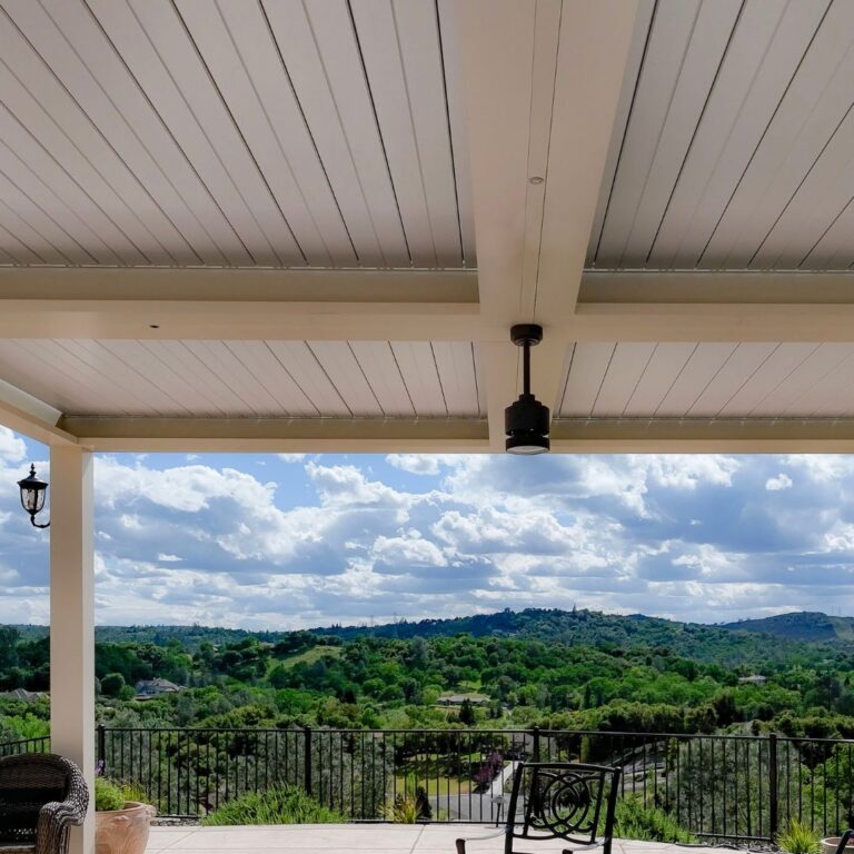 View from beneath a closed white aluminum louvered roof with a compact ceiling fan mounted at center, overlooking a wide valley and rolling hills beyond an iron-railed deck