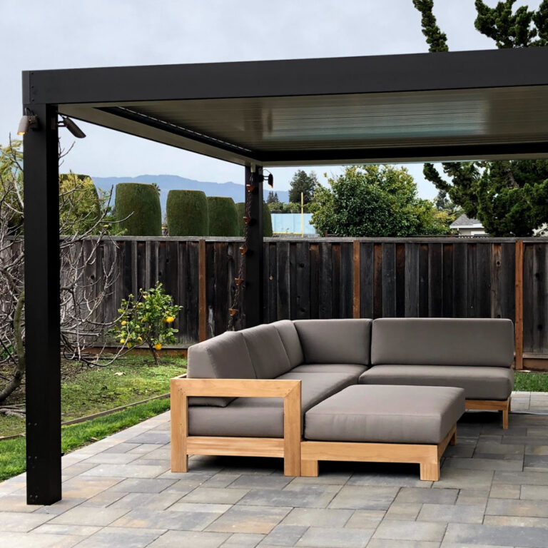 Dark aluminum pergola with closed louvers and a wall-mounted spotlight over a teak sectional sofa on a paver patio with a mountain range visible beyond the fence