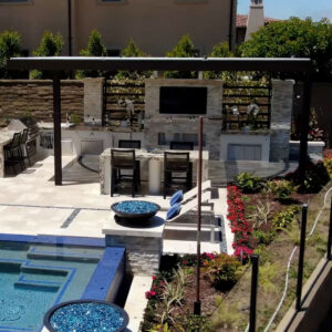 Dark aluminum motorized pergola covering a large outdoor entertaining area with a built-in kitchen, bar seating, wall-mounted TV, fireplace, and pool beyond