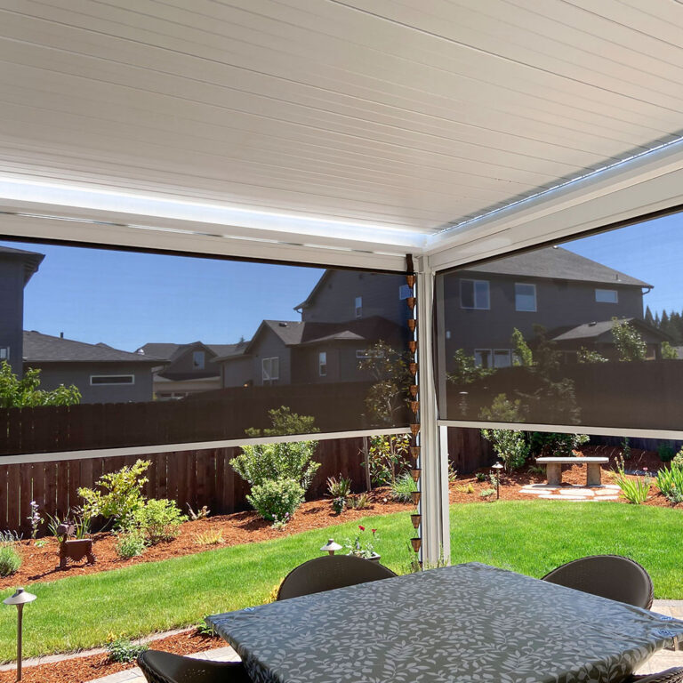 Under-canopy view of a white aluminum louvered pergola with closed louvers, LED perimeter lighting, and two motorized privacy screens fully deployed around a patio dining area