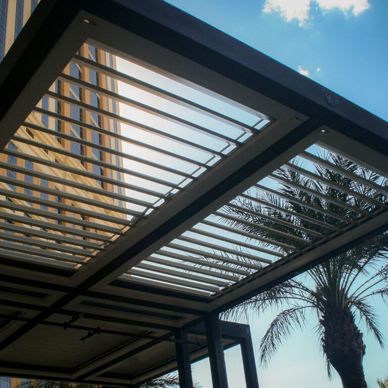 Upward view through fully open aluminum pergola louvers framing a blue sky, palm trees, and a resort tower beyond