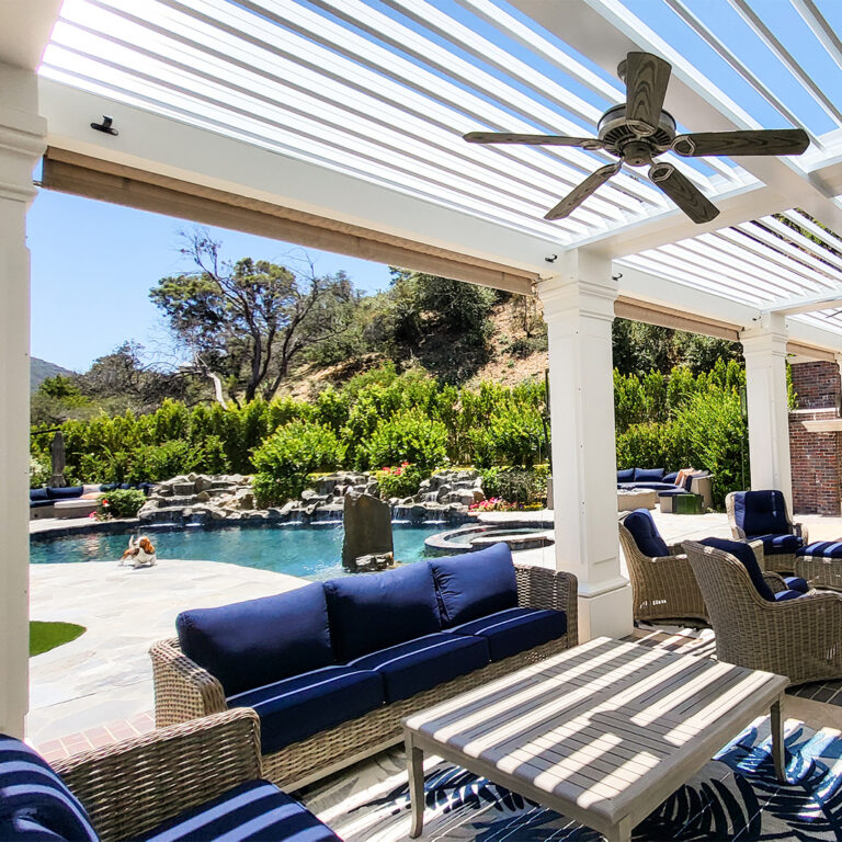White louvered pergola with open louvers and ceiling fan above navy wicker seating overlooking a resort-style pool with a waterfall feature