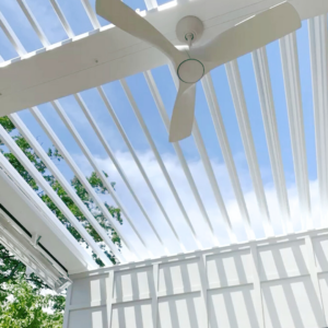 Upward view of fully open white aluminum louvers on a pergola with a three-blade ceiling fan mounted to the frame and motorized side screens partially deployed
