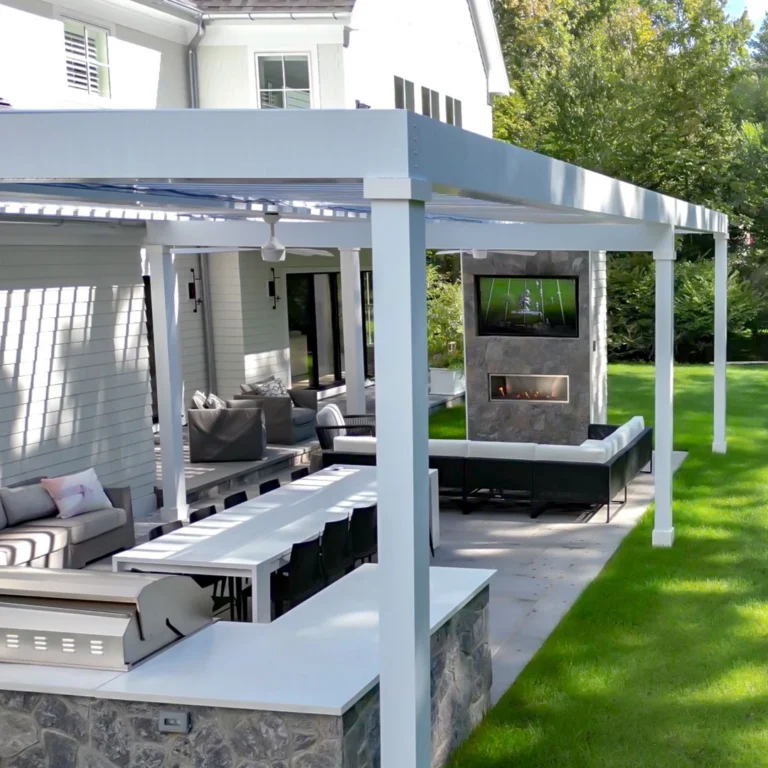 White aluminum louvered pergola covering an outdoor kitchen, dining area, ceiling fan, and stone fireplace with mounted TV beside a suburban home