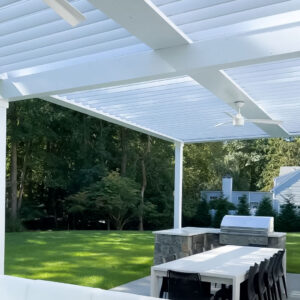 View from beneath a white aluminum louvered roof covering an outdoor kitchen with a stone-clad built-in grill, dining table, and ceiling fan