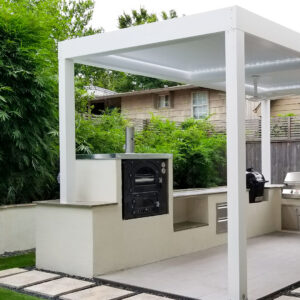 White aluminum louvered pergola with closed louvers over a built-in outdoor kitchen featuring a wood-fired pizza oven, kamado grill, and stainless steel grill station surrounded by bamboo
