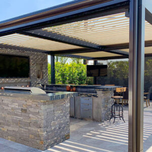 Dark-framed aluminum louvered pergola with open cream louvers casting shadow stripes over a full outdoor kitchen with stacked slate stone surround, granite countertop bar, built-in grill, stainless drawers, and bar stools