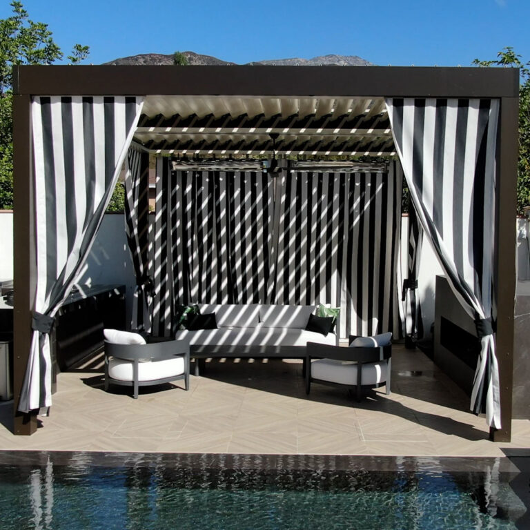 Freestanding dark-framed aluminum louvered pergola with open louvers and floor-length black-and-white striped curtains tied back on all sides, forming a poolside cabana with lounge seating and a mountain backdrop