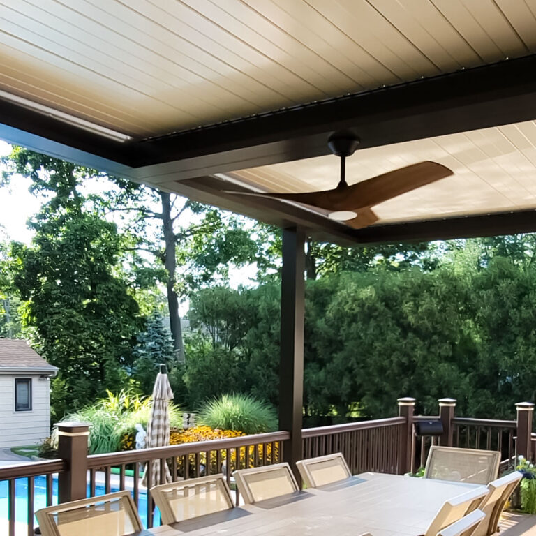 Under-canopy view of a dark-framed aluminum louvered pergola with closed louvers, a wood-blade ceiling fan, integrated lighting, and a pool visible beyond the deck railing