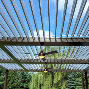 Upward view through fully open white louvers on a multi-bay dark aluminum pergola showing two bronze ceiling fans with light kits against a blue sky with a weeping willow beyond