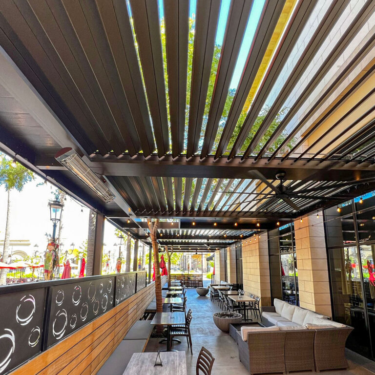 Restaurant pergola interior showing infrared heaters, ceiling fan, and string lights mounted under open aluminum louvers along a long dining patio
