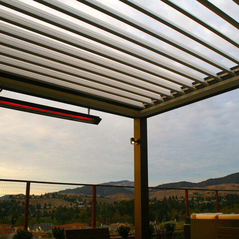 Under-canopy view of a dark aluminum louvered pergola at dusk with a glowing infrared heater mounted to the beam, a post-mounted spotlight, and a wide valley and mountain panorama through cable railings