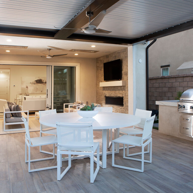 Dark aluminum louvered pergola with closed louvers and dual ceiling fans at dusk covering a round white dining table, stone fireplace with TV, and built-in grill, with open sliding doors to a lit interior