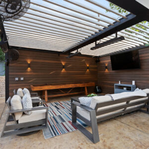 Interior view of a louvered pergola with open louvers, infrared heater, ceiling fan, outdoor TV, and shuffleboard table on a wood slat wall