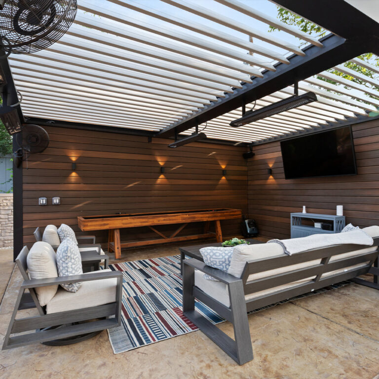 Interior view of a louvered pergola with open louvers, infrared heater, ceiling fan, outdoor TV, and shuffleboard table on a wood slat wall