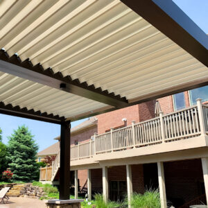 Close-up view from beneath a dark-framed aluminum pergola with fully closed cream louvers beside a multi-level brick home with an upper deck and landscaped yard
