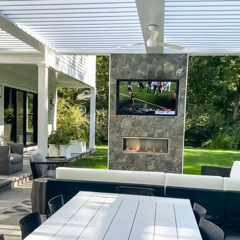 White aluminum louvered pergola with a ceiling fan covering an outdoor dining and lounge area facing a stone fireplace wall with a mounted television playing a football game