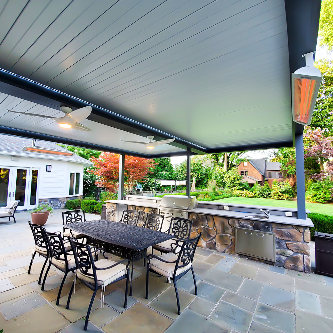 Louvered Pergola Over Outdoor Kitchen With Infrared Heater and Fans Under-canopy view of a dark aluminum louvered pergola with closed louvers, dual ceiling fans, and a glowing wall-mounted infrared heater over a flagstone patio with a cast iron dining set and stone outdoor kitchen