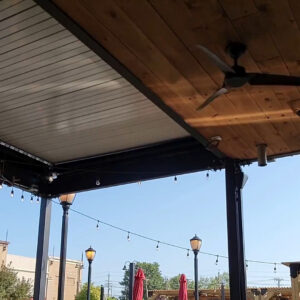 Underside view of a commercial louvered pergola with closed aluminum louvers and an adjacent wood-paneled ceiling with ceiling fan, over a restaurant patio with string lights and street lamps
