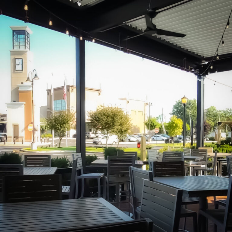 Interior of a louvered pergola patio at Tommy's Tavern with gray dining tables open to a town center street view with clock tower beyond