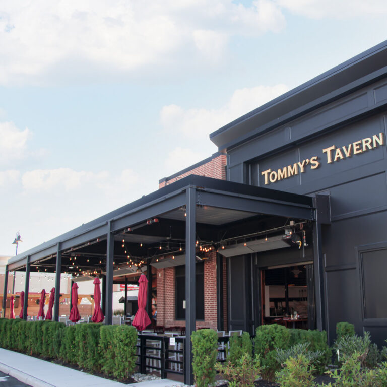 Large closed louvered pergola spanning the full front patio of Tommy's Tavern with mounted heaters, string lights, and dining tables inside