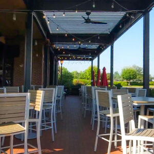 Daytime interior view down a louvered pergola patio at Tommy's Tavern with high-top seating, ceiling fans, string lights, and open tree-line view beyond