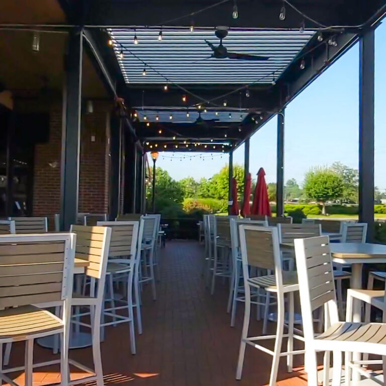 Daytime interior view down a louvered pergola patio at Tommy's Tavern with high-top seating, ceiling fans, string lights, and open tree-line view beyond