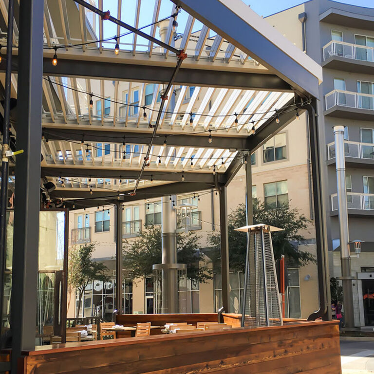 Light aluminum louvered pergola with open louvers over a wood-enclosed restaurant patio with string lights and a freestanding tower heater