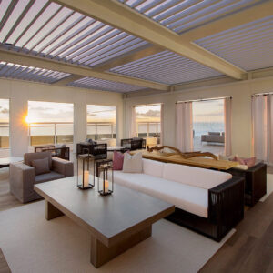 A louvered roof system spans an upscale resort lounge where candlelight and plush seating frame a sweeping waterfront sunset