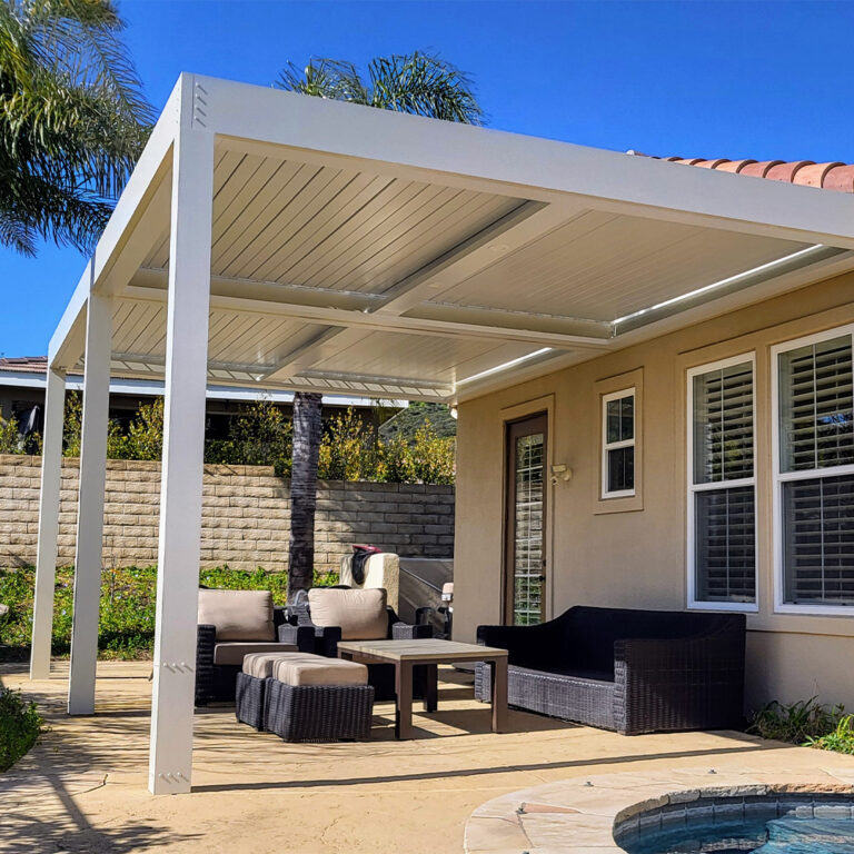 White aluminum louvered pergola with integrated LED lighting attached to a stucco home over a wicker lounge seating area beside a pool