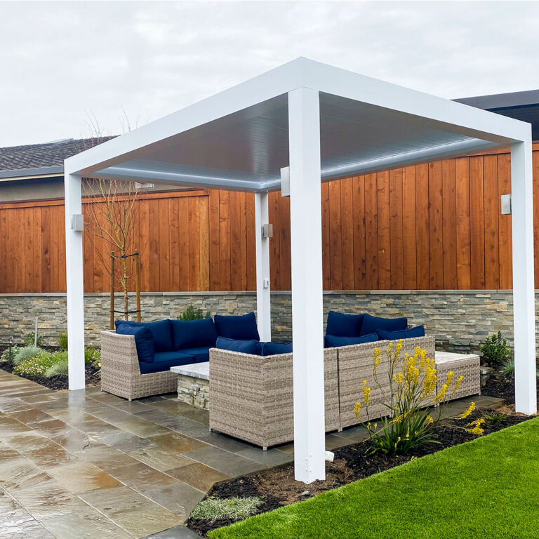 Freestanding white aluminum louvered pergola with closed roof covering a rattan sectional with navy cushions in a landscaped backyard