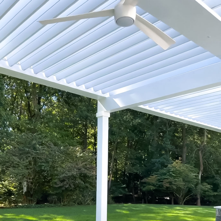 Underside view of a white aluminum louvered pergola with open louvers and a white ceiling fan, framing a lush wooded backyard through the open sides