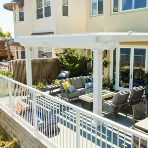 White aluminum louvered pergola with closed louvers attached to a two-story coastal home over an upper deck lounge with a fire table, seating, and a pool visible below the railing