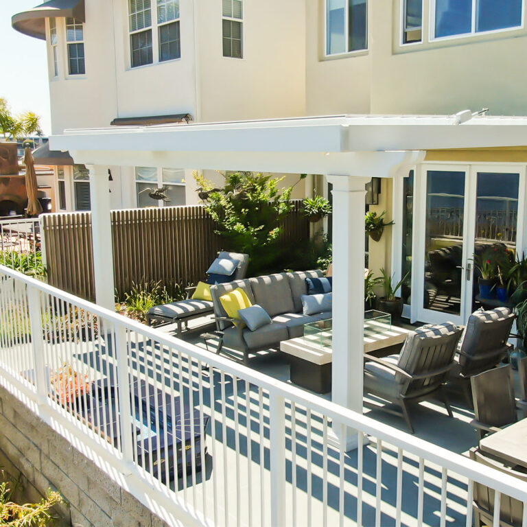 White aluminum louvered pergola with closed louvers attached to a two-story coastal home over an upper deck lounge with a fire table, seating, and a pool visible below the railing