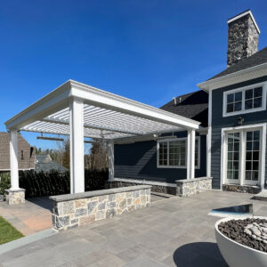 Large white aluminum louvered pergola with open louvers and decorative columns attached to a dark blue colonial-style home, with a stone patio and fire bowl in the foreground