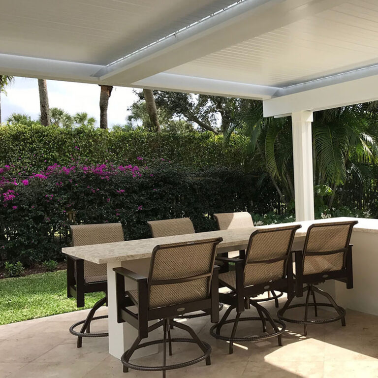 Under-canopy view of a white aluminum louvered pergola with closed louvers and LED perimeter lighting over a travertine patio dining bar with swivel chairs, bougainvillea hedge, and tropical palms beyond