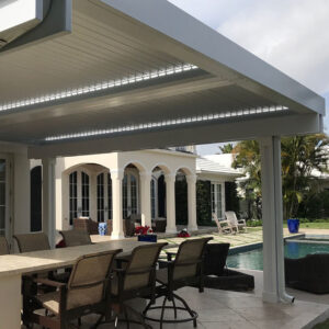 Under-canopy view of a white aluminum louvered pergola with bright LED perimeter lighting and partially open louvers over a dining bar and pool patio, with a tropical estate home with arched loggia and palm trees beyond