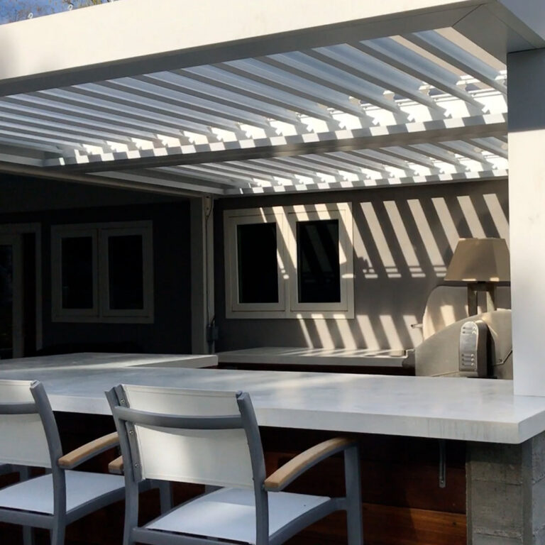 Under-canopy view of a white aluminum louvered pergola with open louvers casting sharp shadow stripes across a modern home wall, above a concrete outdoor bar counter with bar stools and a built-in grill